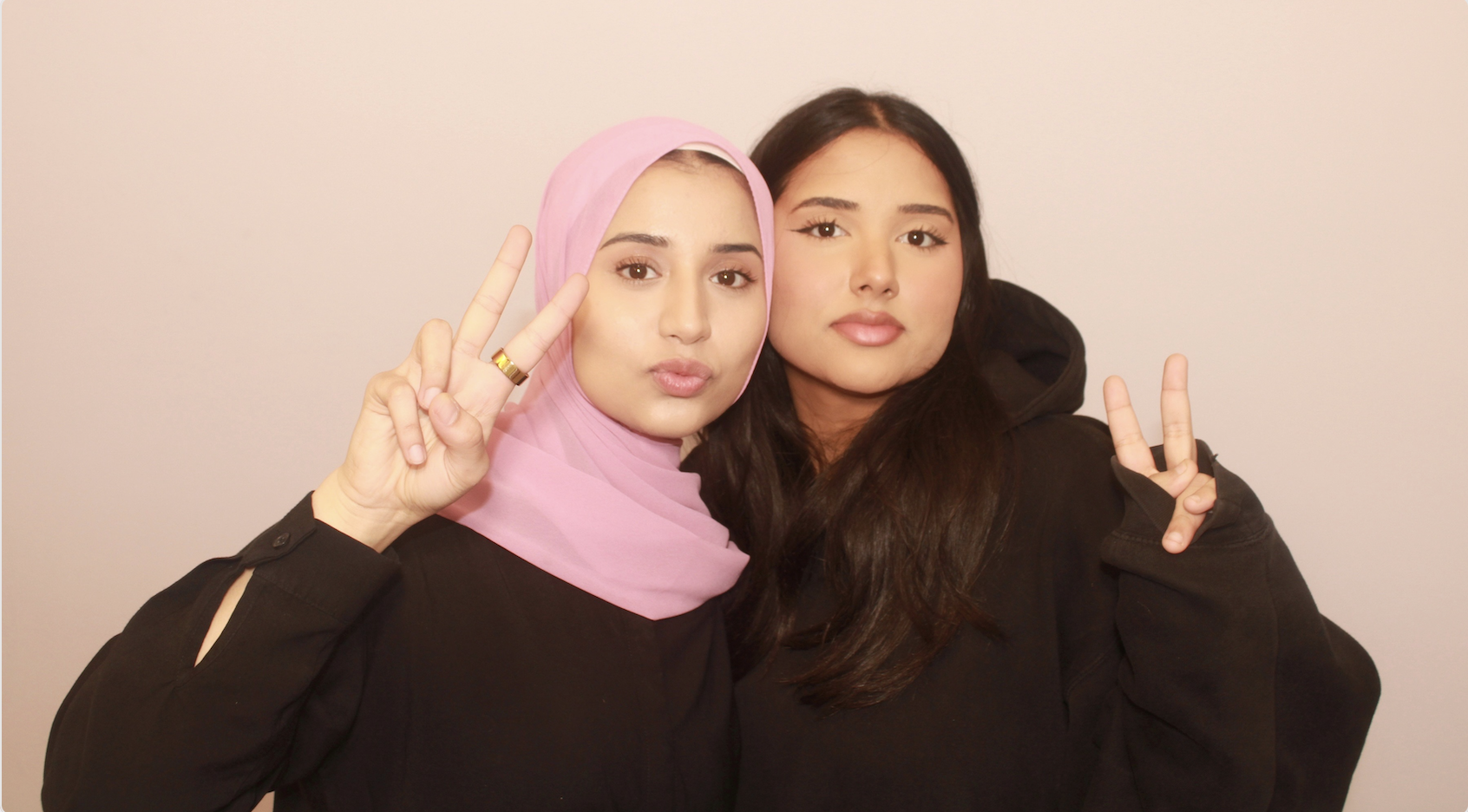 The two sisters behind Mona's Booth Toronto smiling and making peace signs in front of a blush backdrop