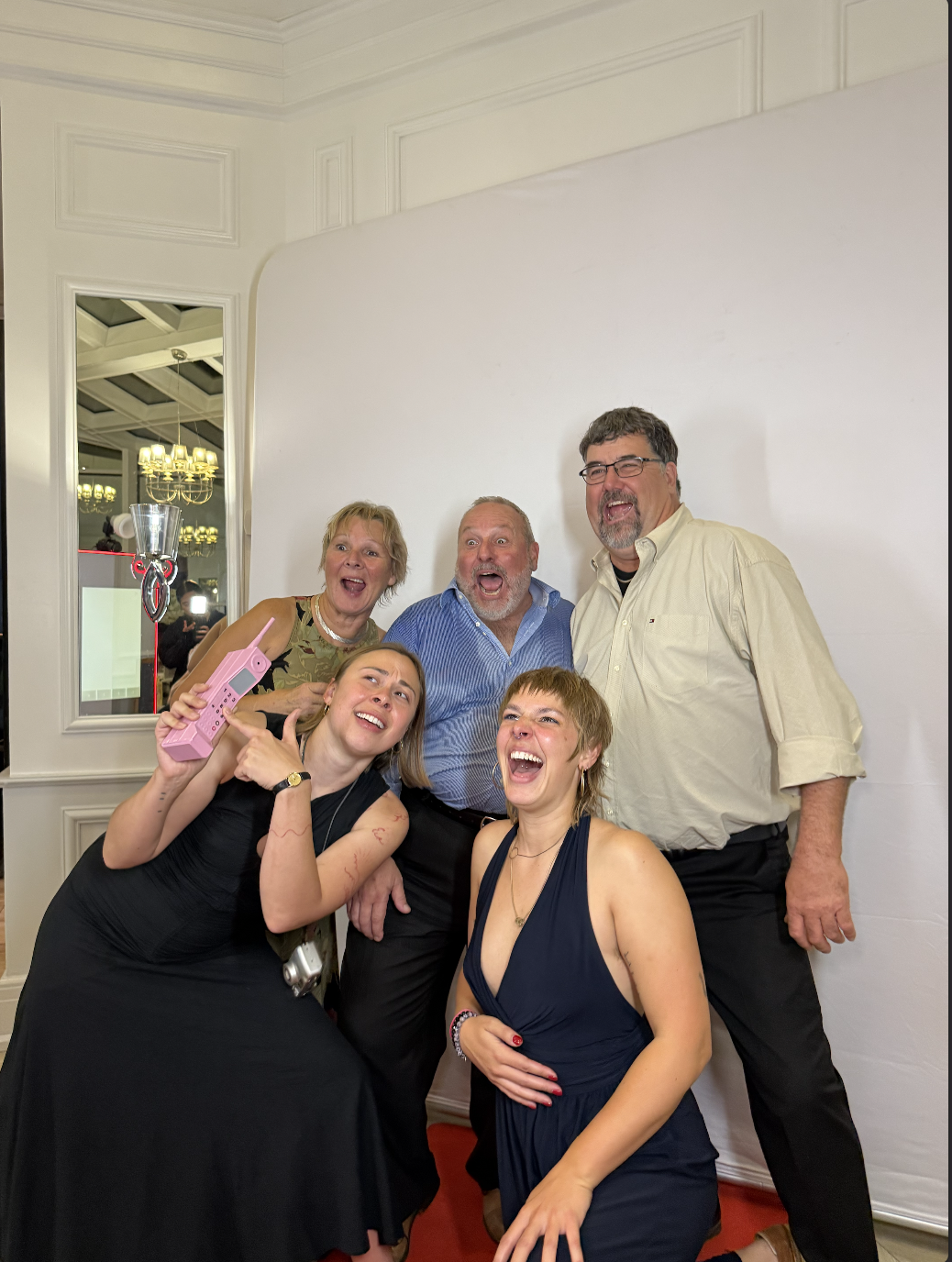 Five guests laughing in front of a white photo booth backdrop, one holding a pink phone prop