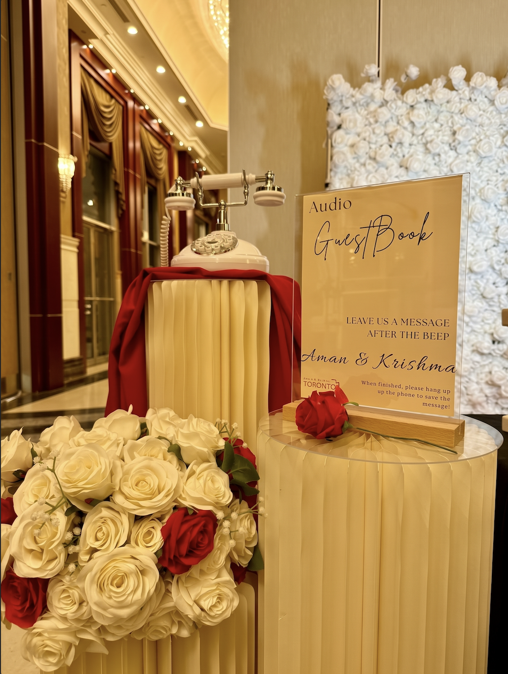 Vintage white audio guestbook telephone on a red-draped pedestal beside a personalized Aman & Krishma sign and roses