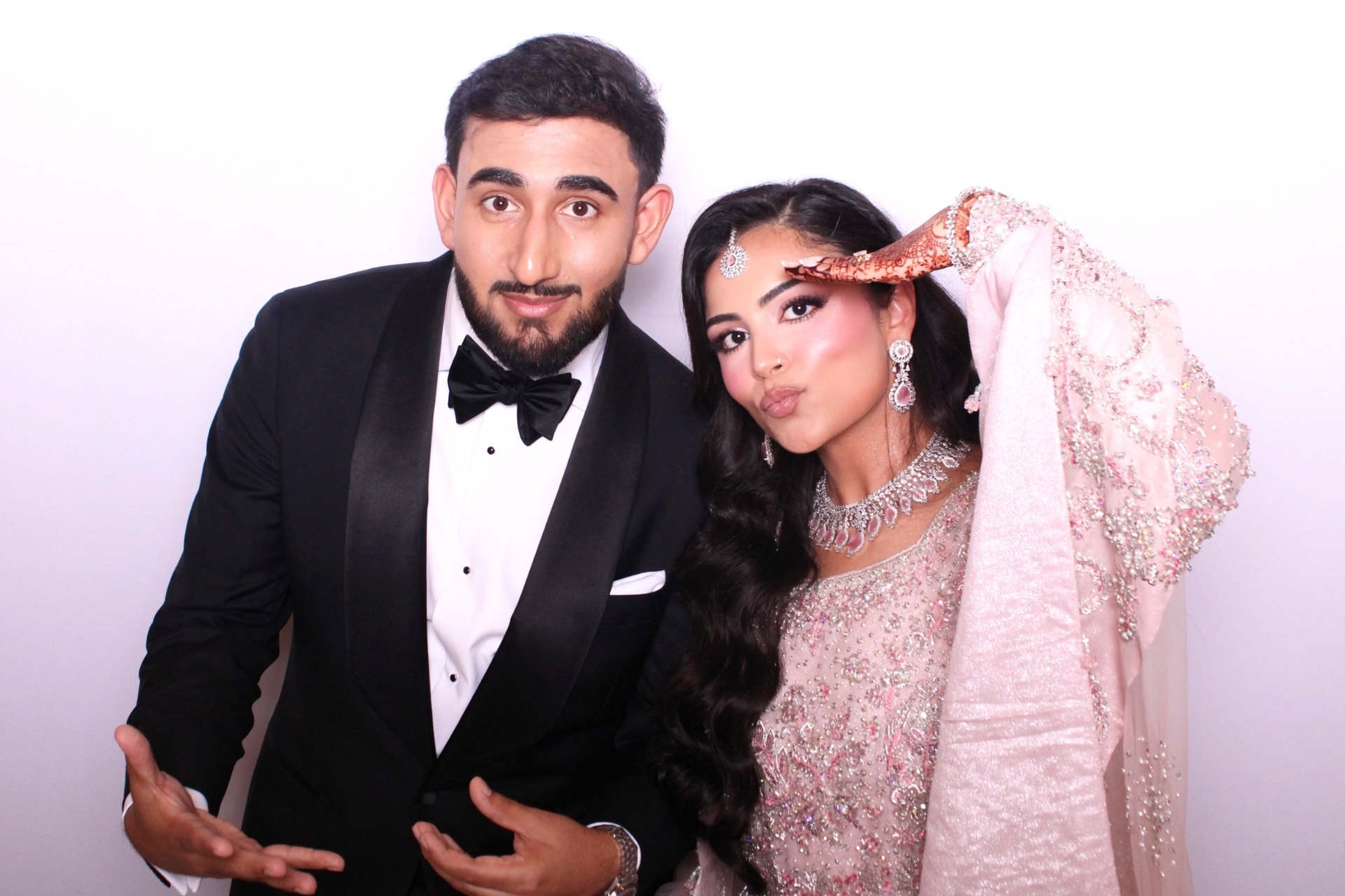Bride in a pink embellished outfit and groom in a tuxedo posing playfully at the Mona's Booth photo booth