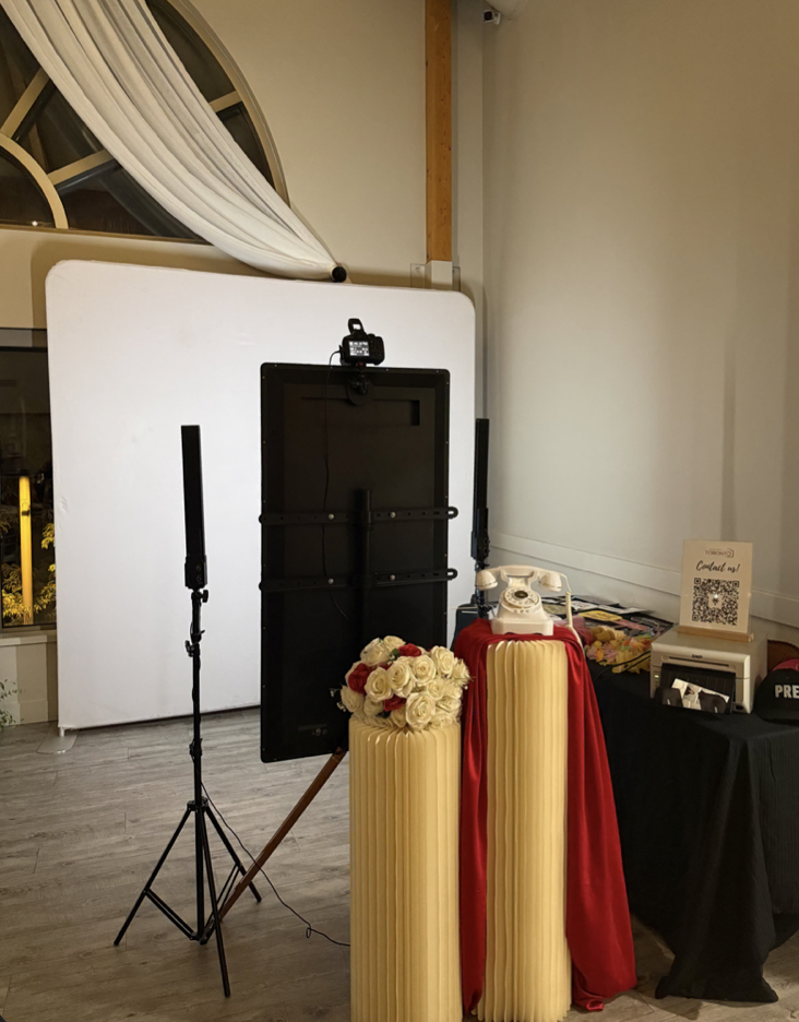 Mona's Booth mirror photo booth set up with a white backdrop, vintage telephone, rose arrangement and print station