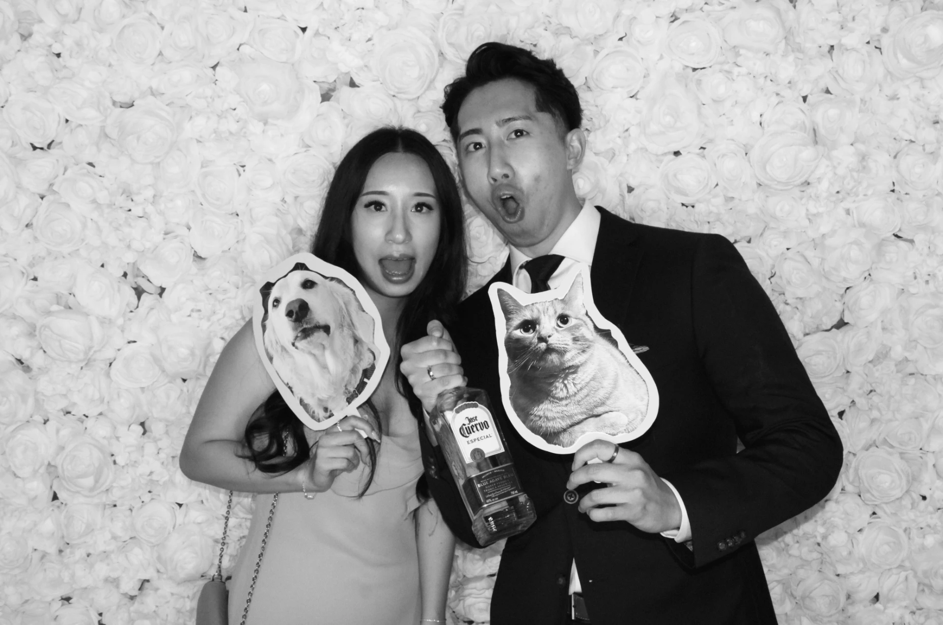 Couple at a white flower wall holding custom dog and cat face props, in black and white