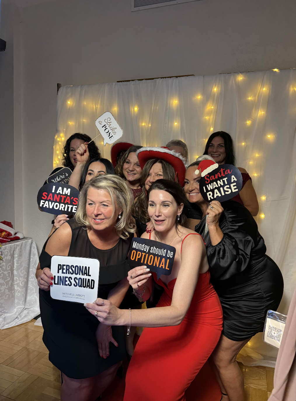 Coworkers holding holiday and branded signs in front of a fairy lights backdrop at a corporate party