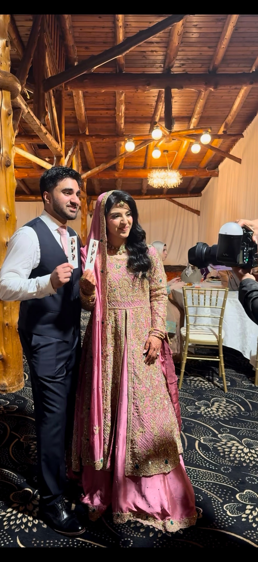 Bride in a pink embellished outfit and groom in a vest each holding up their Mona's Booth photo strip