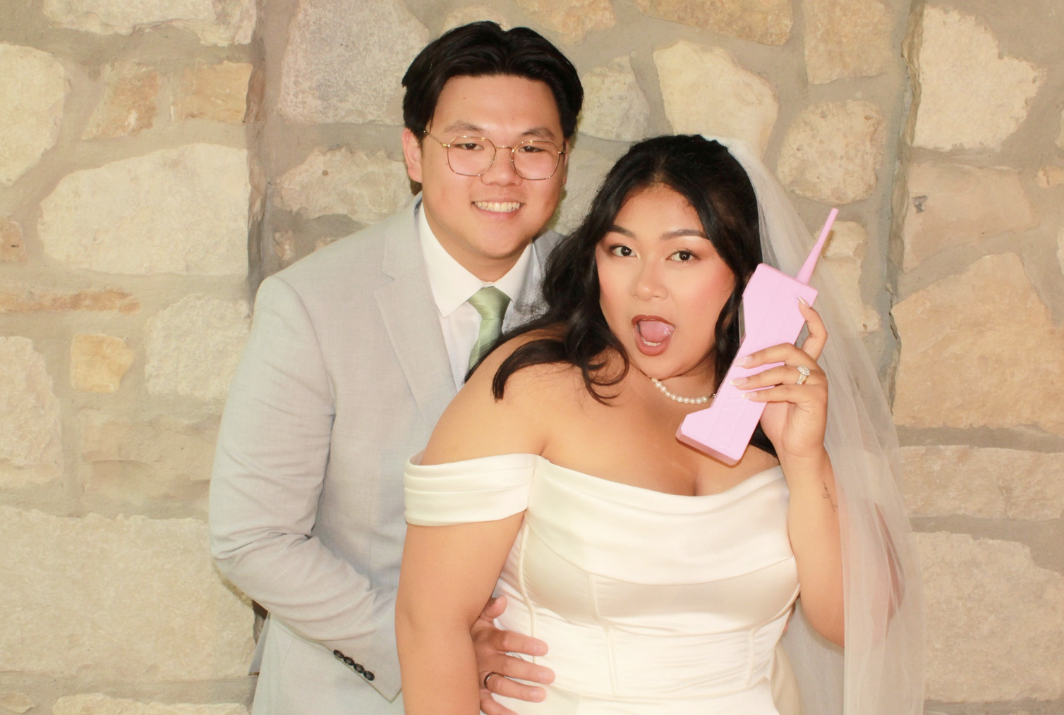 Bride holding a pink retro phone prop beside the groom in front of a stone wall